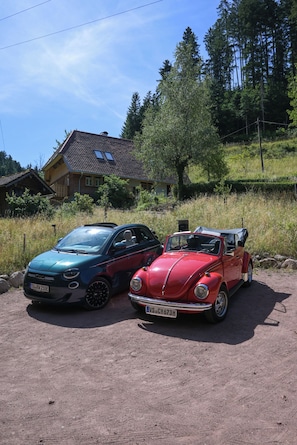 Two vintage cars parked on a dirt surface with a house and trees in the background.