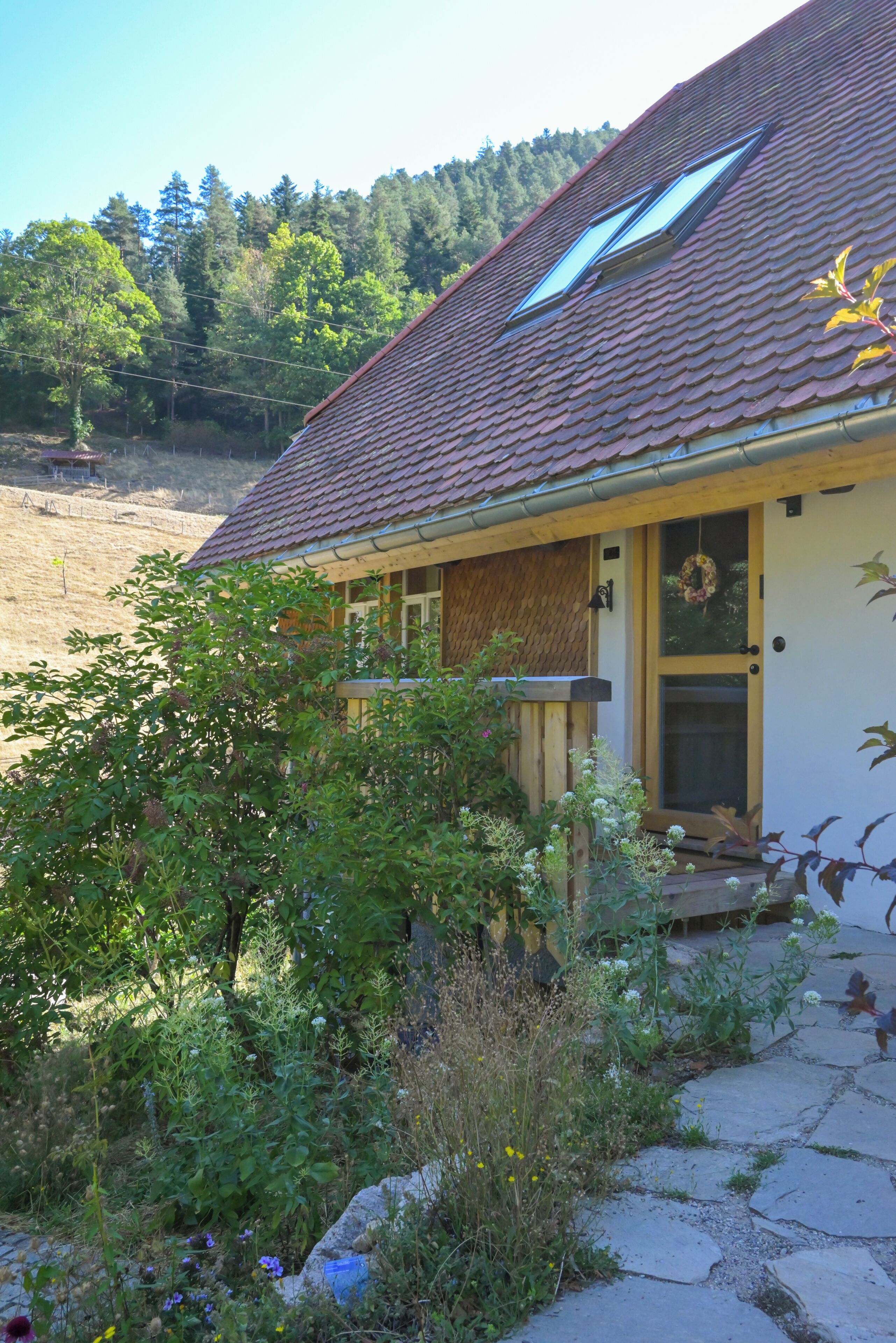 Una casa con tetto spiovente, veranda e un giardino con diverse piante e fiori.