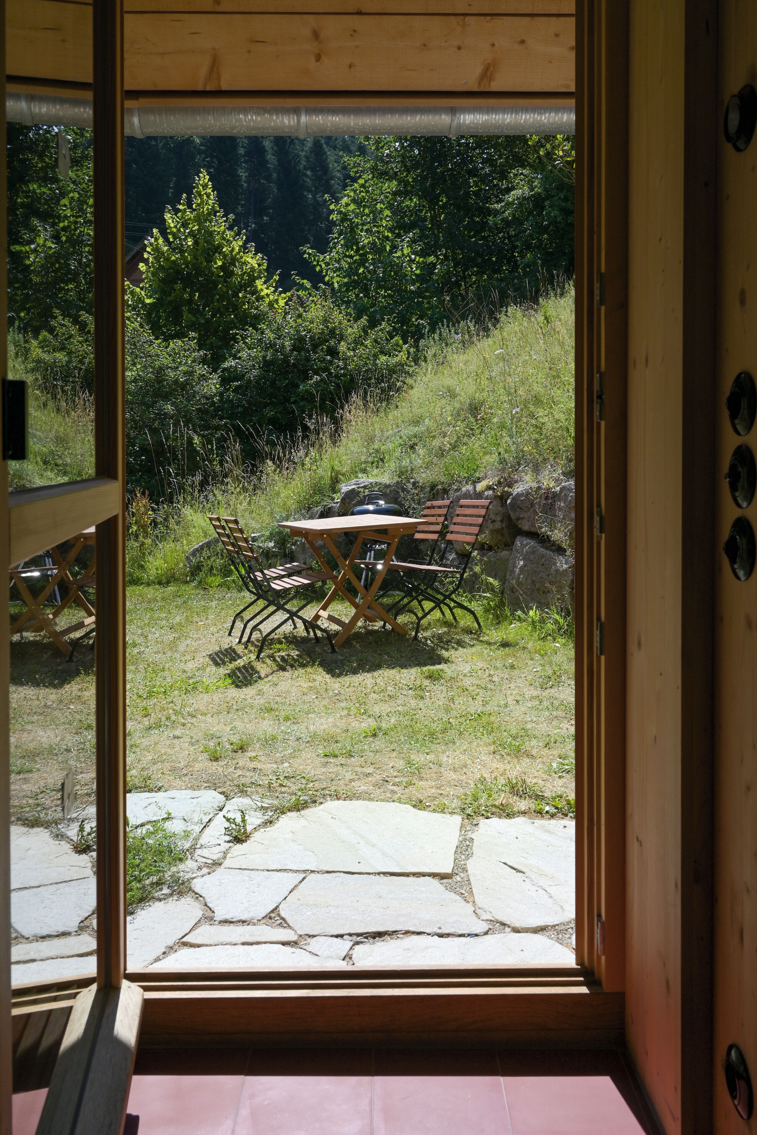 Una finestra con cornice in legno che si affaccia su un'area esterna con tavoli e sedie, immersa nel verde e con un vialetto in pietra.