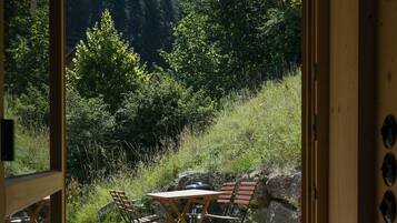 Una finestra con cornice in legno che si affaccia su un'area esterna con tavoli e sedie, immersa nel verde e con un vialetto in pietra.