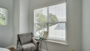 Ein Zimmer mit grauem Sessel, einem kleinen Tisch mit Vase und Bilderrahmen sowie einem Fenster mit Jalousien.