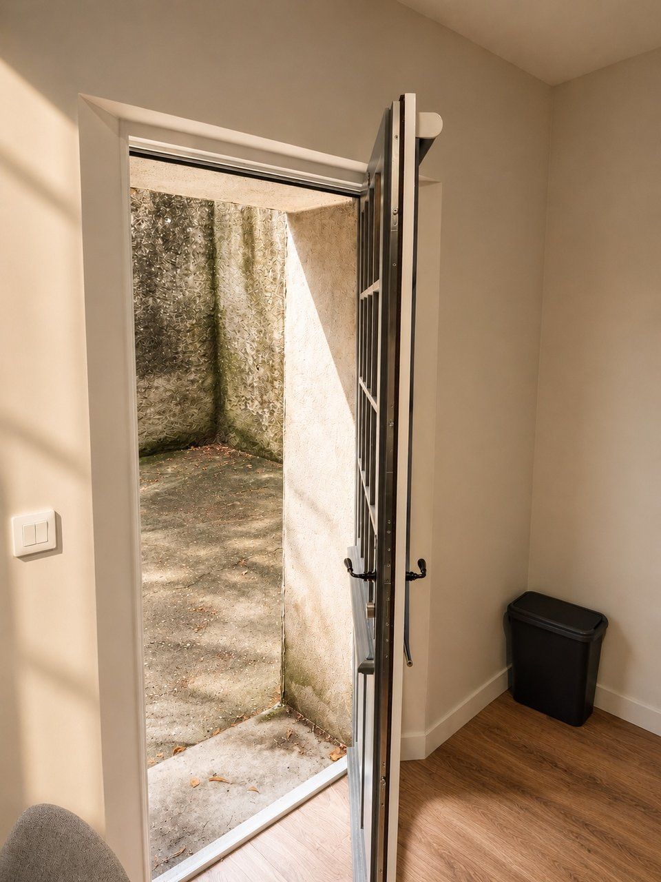 Une pièce avec une porte donnant sur un petit espace extérieur doté d’un sol en béton et d’une poubelle.