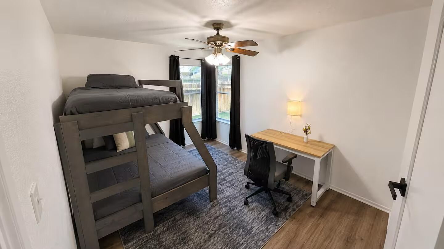 A compact room with a bunk bed, a desk, a chair, a ceiling fan, and a window with curtains.
