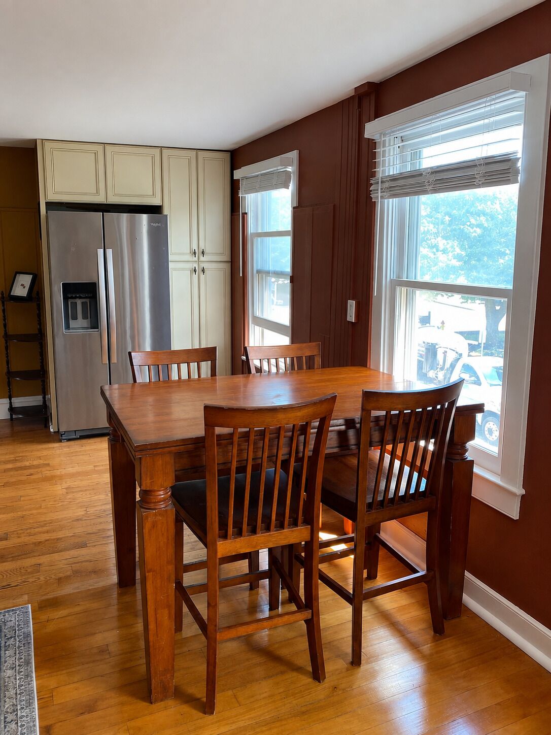 A dining area with a wooden table and chairs, a stainless steel refrigerator, and a window with blinds.