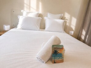 Ein ordentlich bezogenes Bett mit weißen Leinen und Kissen, ein gefaltetes Handtuch und ein Willkommen-Schild mit einer Palme und Sonnenuntergang.