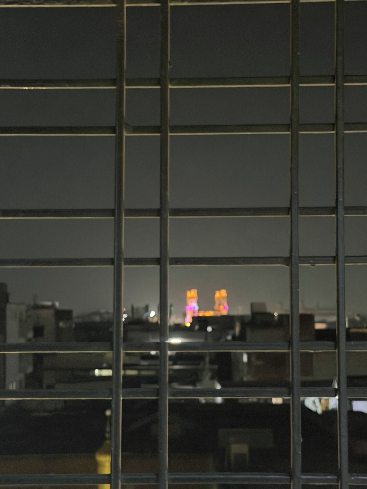 Vista a través de una ventana con persianas horizontales, mostrando un paisaje urbano con edificios iluminados a lo lejos.
