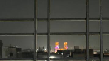 Vista a través de una ventana con persianas horizontales, mostrando un paisaje urbano con edificios iluminados a lo lejos.