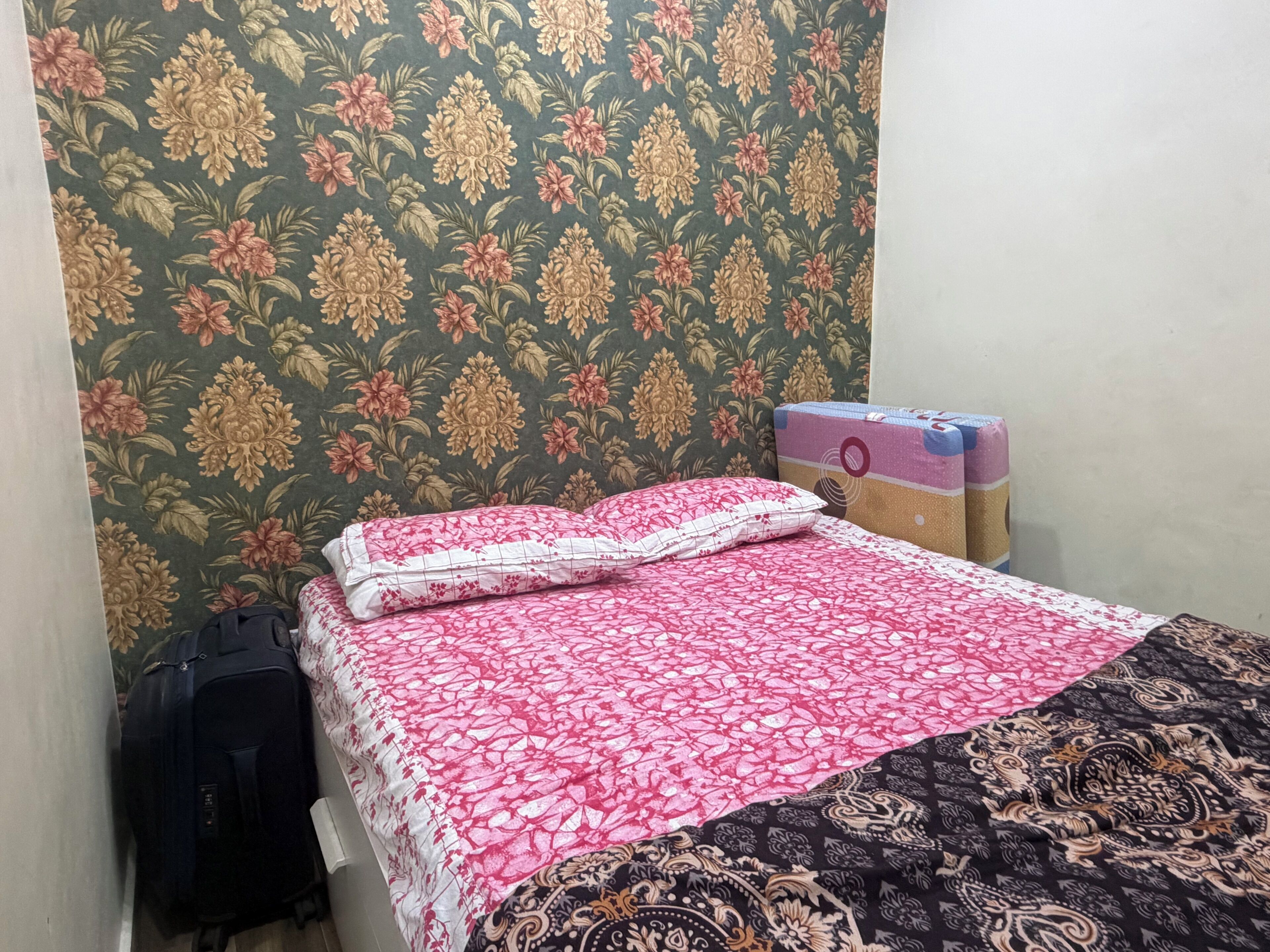 Een bed met een bloemenprintdeken en een kussen met dessin, een koffer en een doos tegen een behangwand.