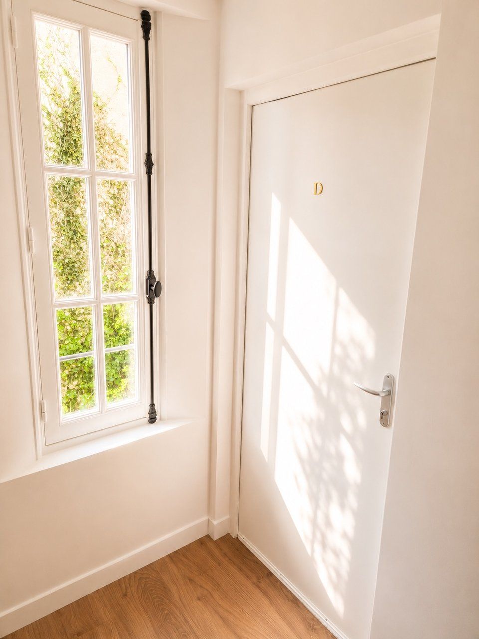 Una puerta blanca con una pequeña 'D' dorada, una ventana con cortina de madera y un piso de madera.