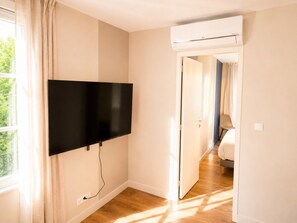 Een kamer met een wandgemonteerde flatscreen televisie, een airconditioningunit boven de deur, en uitzicht op de aangrenzende kamer door een geopende deur.