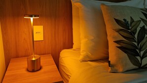 Ein ordentlich bezogenes Bett mit einem Kopfkissen mit Blättermuster, ein Nachttisch mit einer Lampe und eine Steckdose.