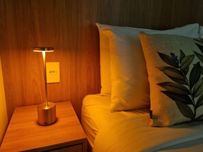 Un llit ben fet amb un coixí amb un dibuix de fulles, una taula de nit amb una làmpada i una presa elèctrica a la paret.