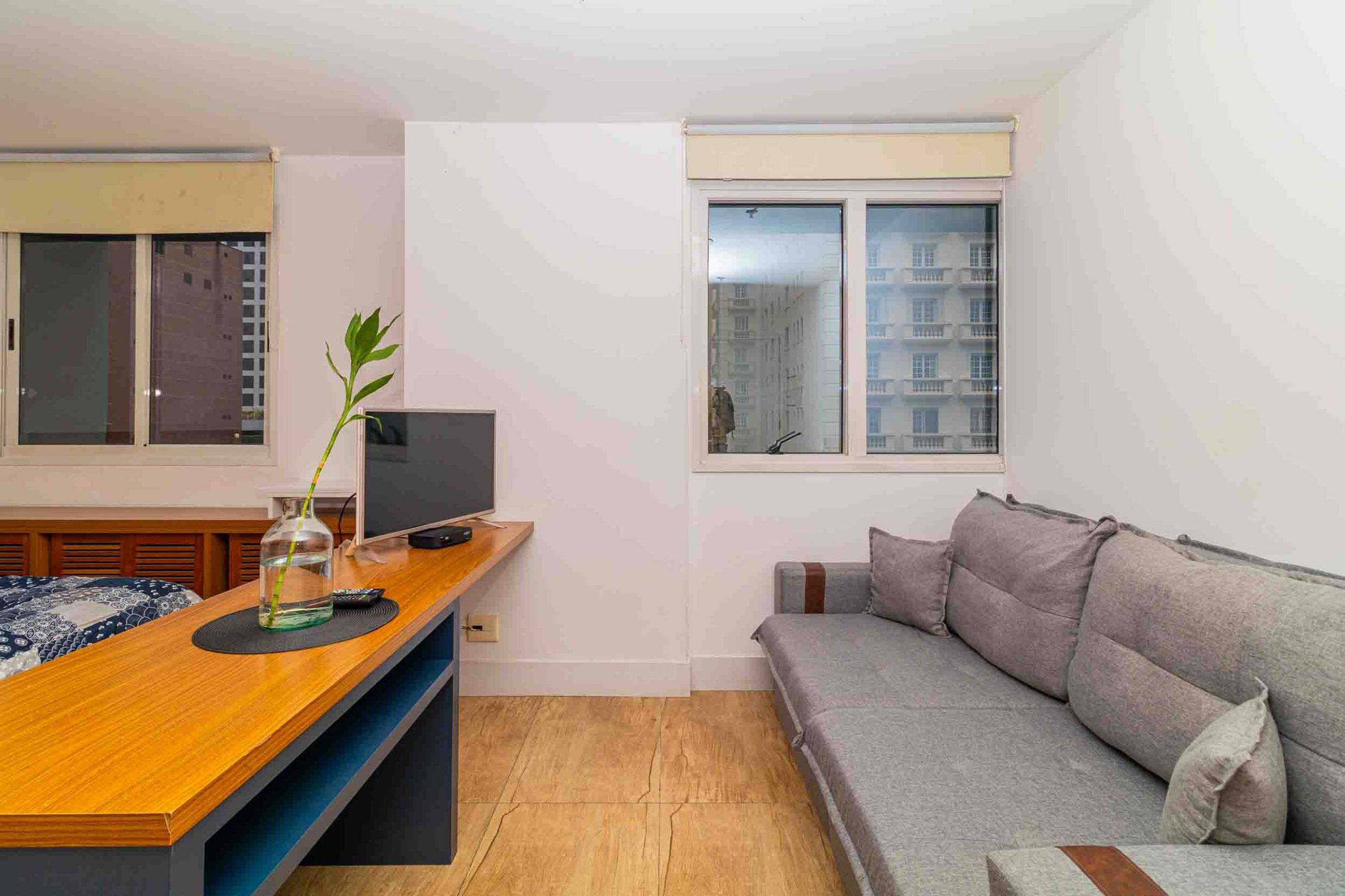 A modern living room with a wooden coffee table, a grey sofa, a television, and a window with a view of buildings.