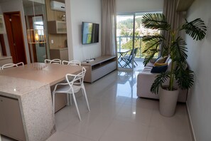 A modern living room with a dining area, a flat-screen TV mounted on the wall, and a large potted plant. - Apto com Garagem a 600m da Praia das Toninhas (Ubatuba)