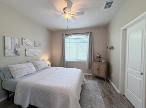 Ein Schlafzimmer mit Bett, Deckenventilator, Fenster mit Vorhängen und Wandbildern.