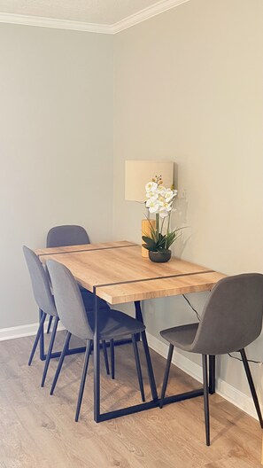 A dining area with a wooden table, four chairs, a potted plant, and a wall-mounted lamp. - The Cove #2. Lovely 2-bedroom apartment with AC in charming Rockingham. (Rockingham)