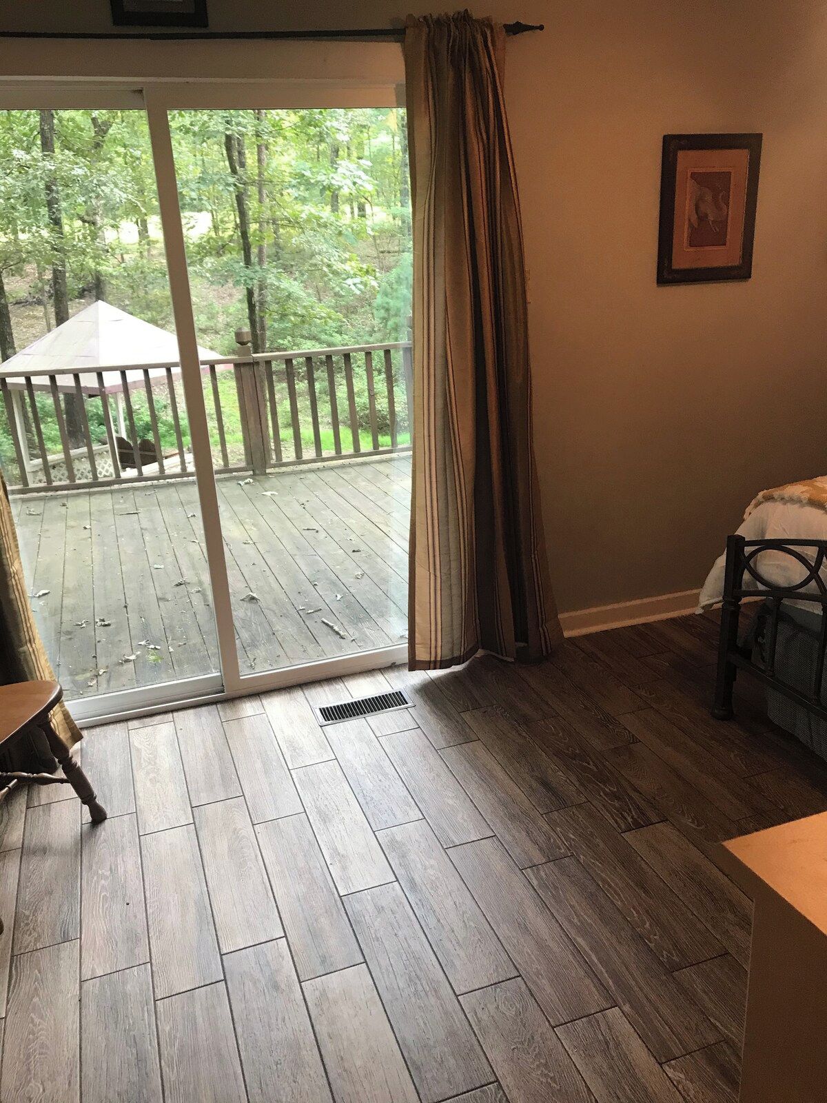 A room with a wooden floor, a sliding glass door leading to an outdoor deck, a chair, and a bed.