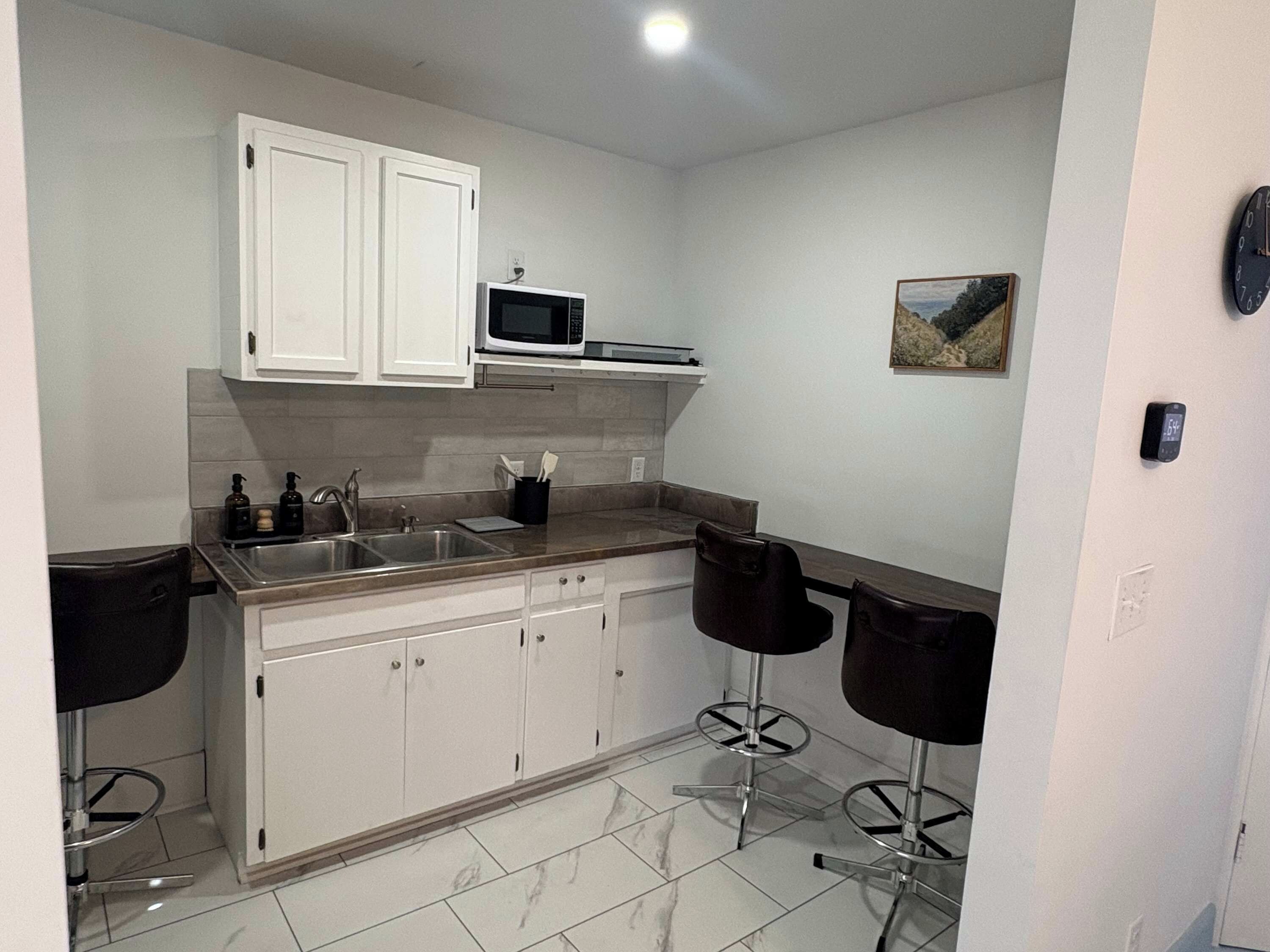 A compact kitchen with white cabinets, a microwave, a sink, and two bar stools.