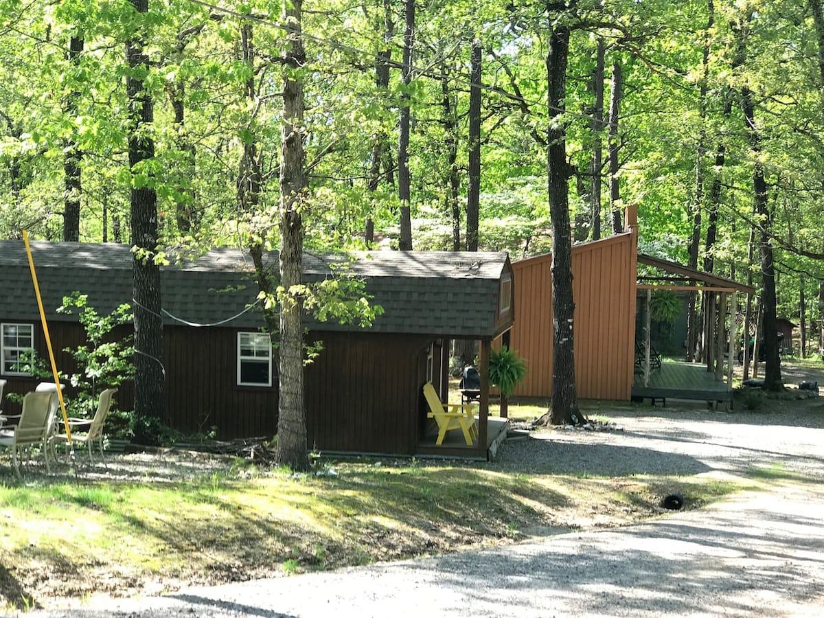 A cabin with a porch, a small deck, and a covered outdoor seating area surrounded by trees.