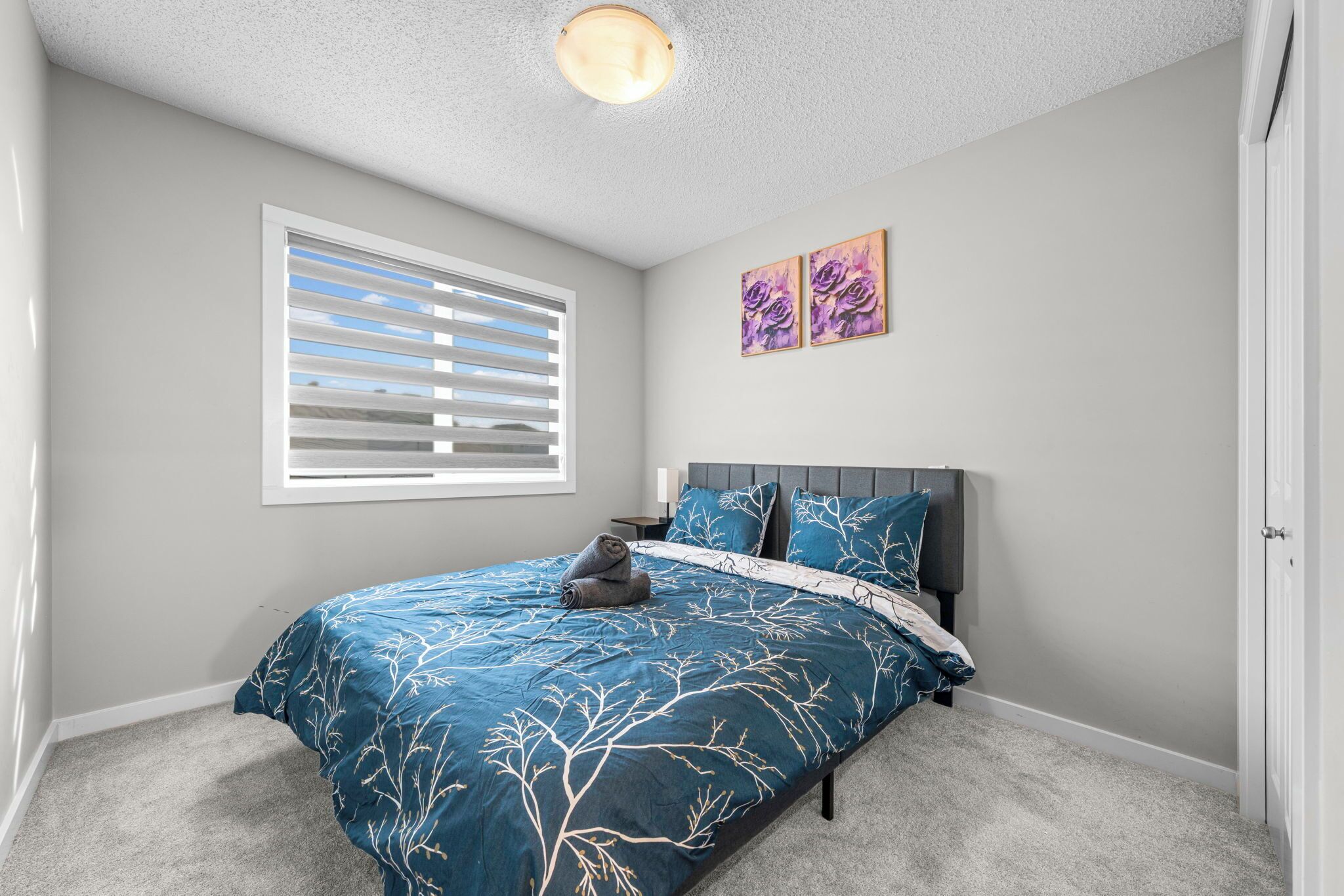 A bedroom with a bed, a window with blinds, a ceiling light, and two wall-mounted paintings.