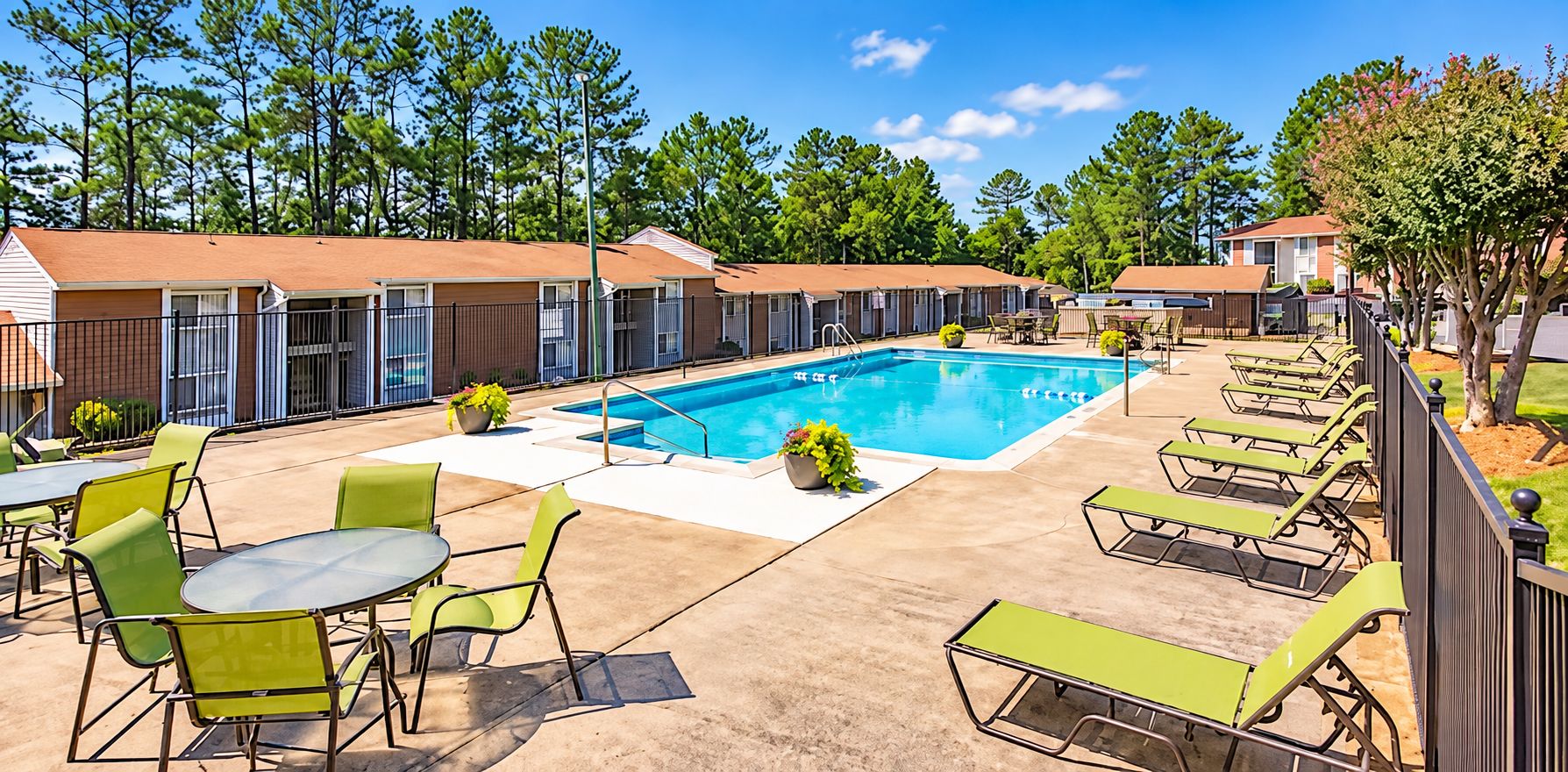 A swimming pool area with lounge chairs and tables, surrounded by a fence and trees.