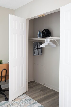 Une pièce avec une porte blanche, une petite table sur laquelle se trouve une plante, une chaise, un étendoir suspendu et un ventilateur fixé au mur.