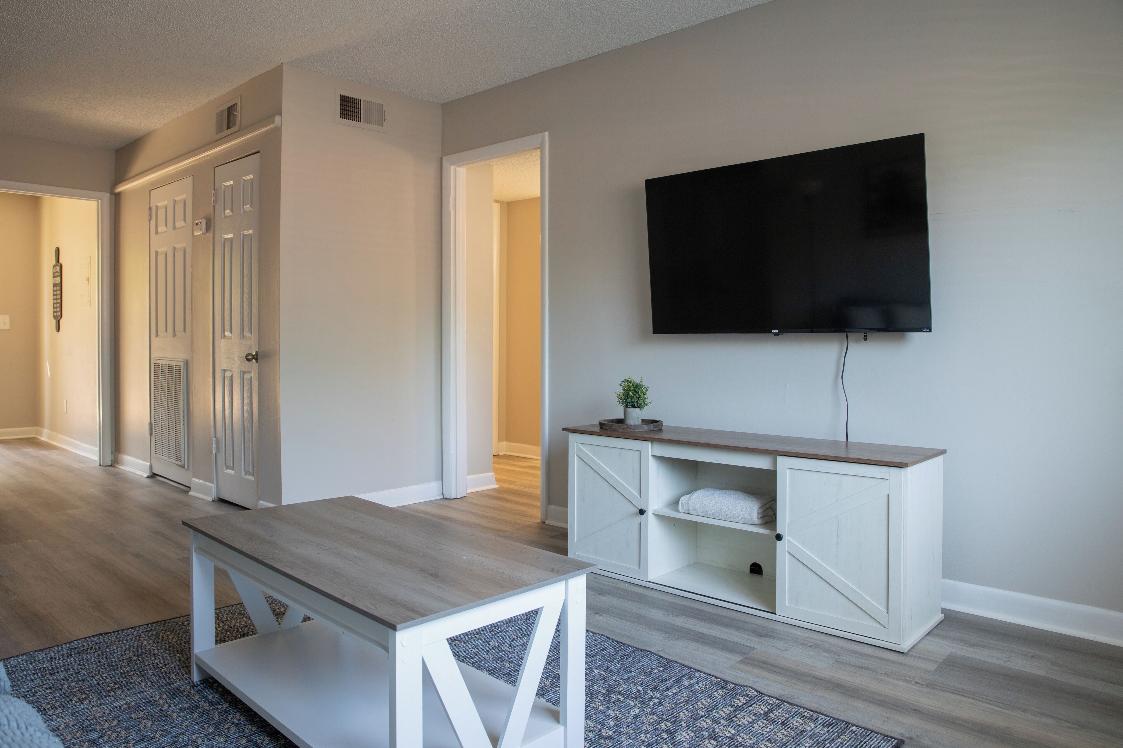 Un salon moderne avec une table basse en bois, un meuble de rangement blanc intégrant un téléviseur à écran plat, et un couloir menant à une autre pièce.