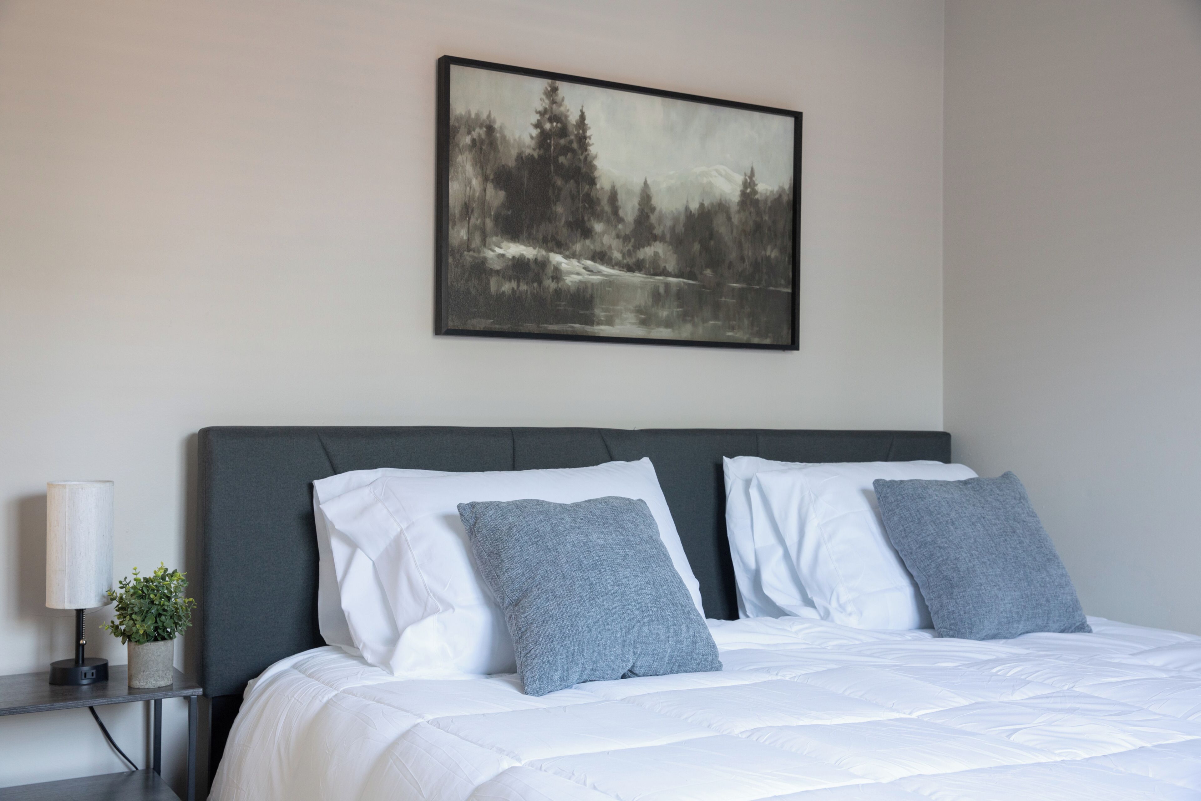 Une chambre à coucher avec un lit, des oreillers, une table de chevet avec une lampe et un tableau encadré au mur.