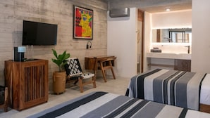 Quarto moderno com cama, escrivaninha, cadeira, televisão e banheiro visível através de uma porta aberta.