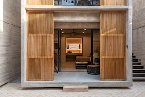 A modern building with a concrete facade, wooden slatted doors, and a visible interior with a sofa and a dining area.