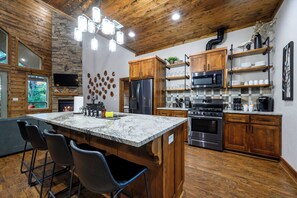 A modern kitchen with a large island, wooden cabinets, a stone fireplace, and a stone wall. - Secluded Cabin | Hot Tub | Game Loft (Broken Bow)