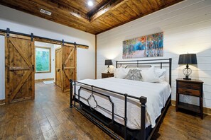 A bedroom with a large bed, wooden flooring, a painting on the wall, and a sliding barn door. - Secluded Cabin | Hot Tub | Game Loft (Broken Bow)