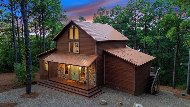 A two-story log cabin with a stone foundation, a covered porch, and a metal roof, surrounded by a forest. - Secluded Cabin | Hot Tub | Game Loft (Broken Bow)