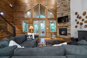 Ein Holzblockhaus-Interieur mit Kamin, einem an einer Steinwand montierten Fernseher und einer gemütlichen Sitzecke mit Sofa und Couchtisch.