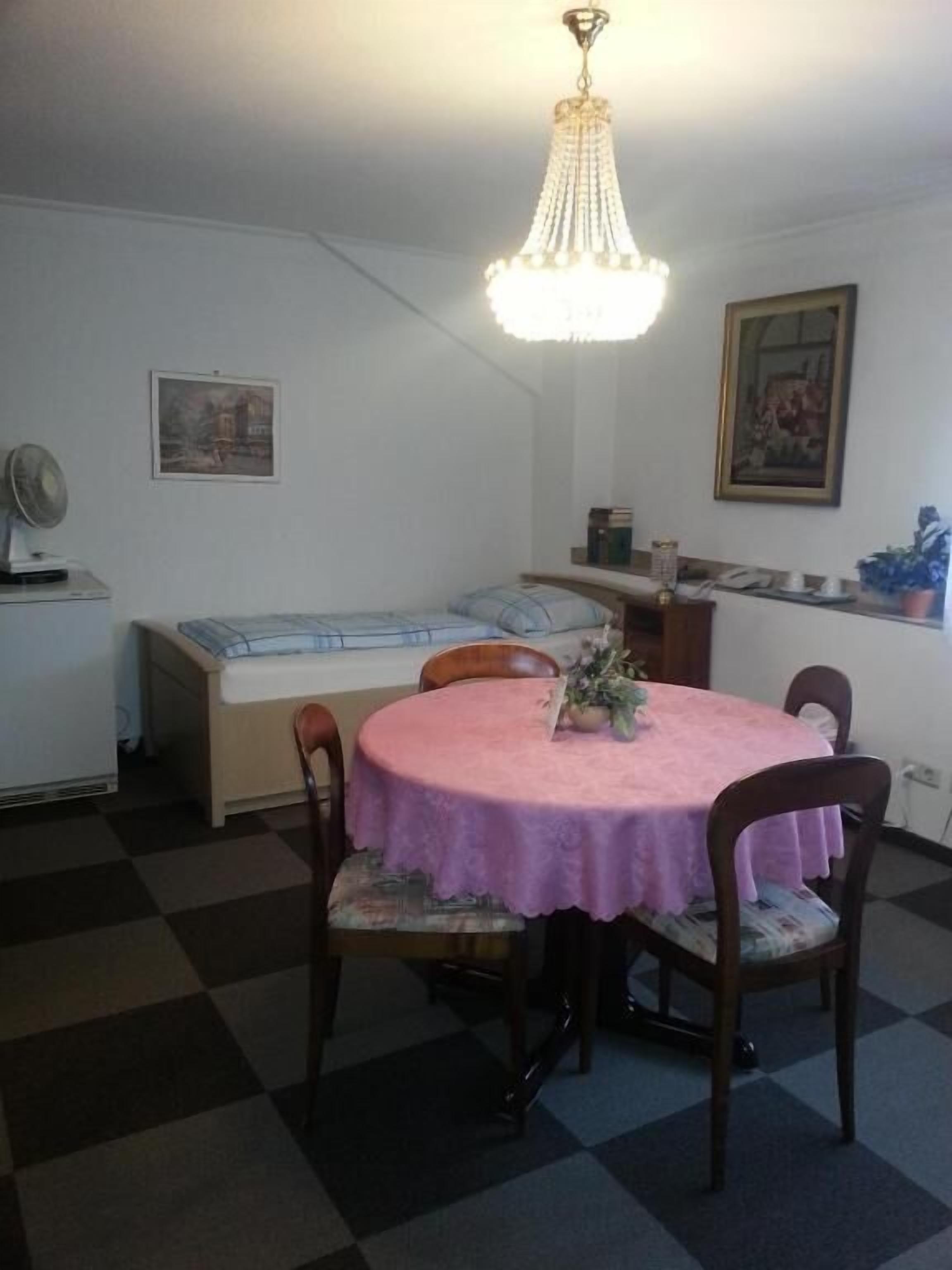 Ein Essbereich mit einem runden Tisch, der mit einer rosa Tischdecke bedeckt ist, umgeben von Holzsesseln. Eine Kristallleuchter hängt von der Decke, und es befinden sich zwei gerahmte Bilder an den Wänden.