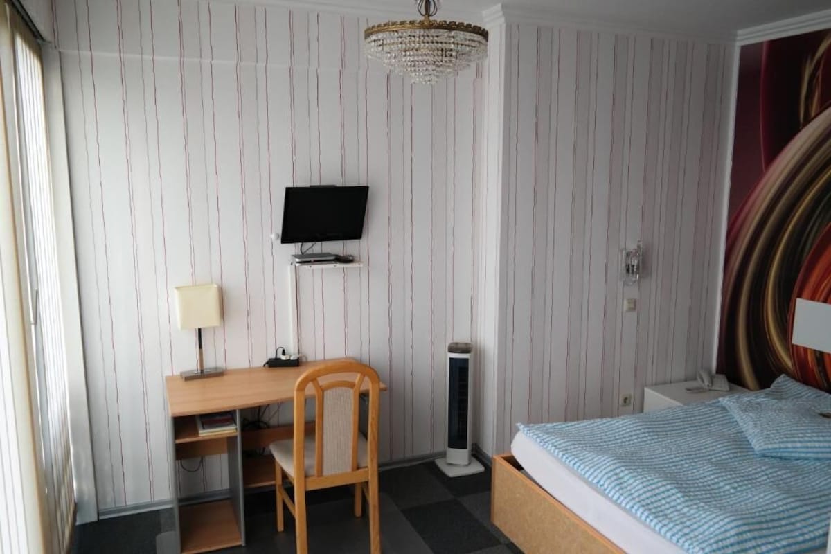 A hotel room with a bed, a desk with a chair, a television, a standing heater, and a chandelier.