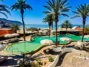 A resort pool area with lounge chairs, umbrellas, and palm trees. - Puerta Cortés 404 - 2BD Marina View Condo (La Paz)