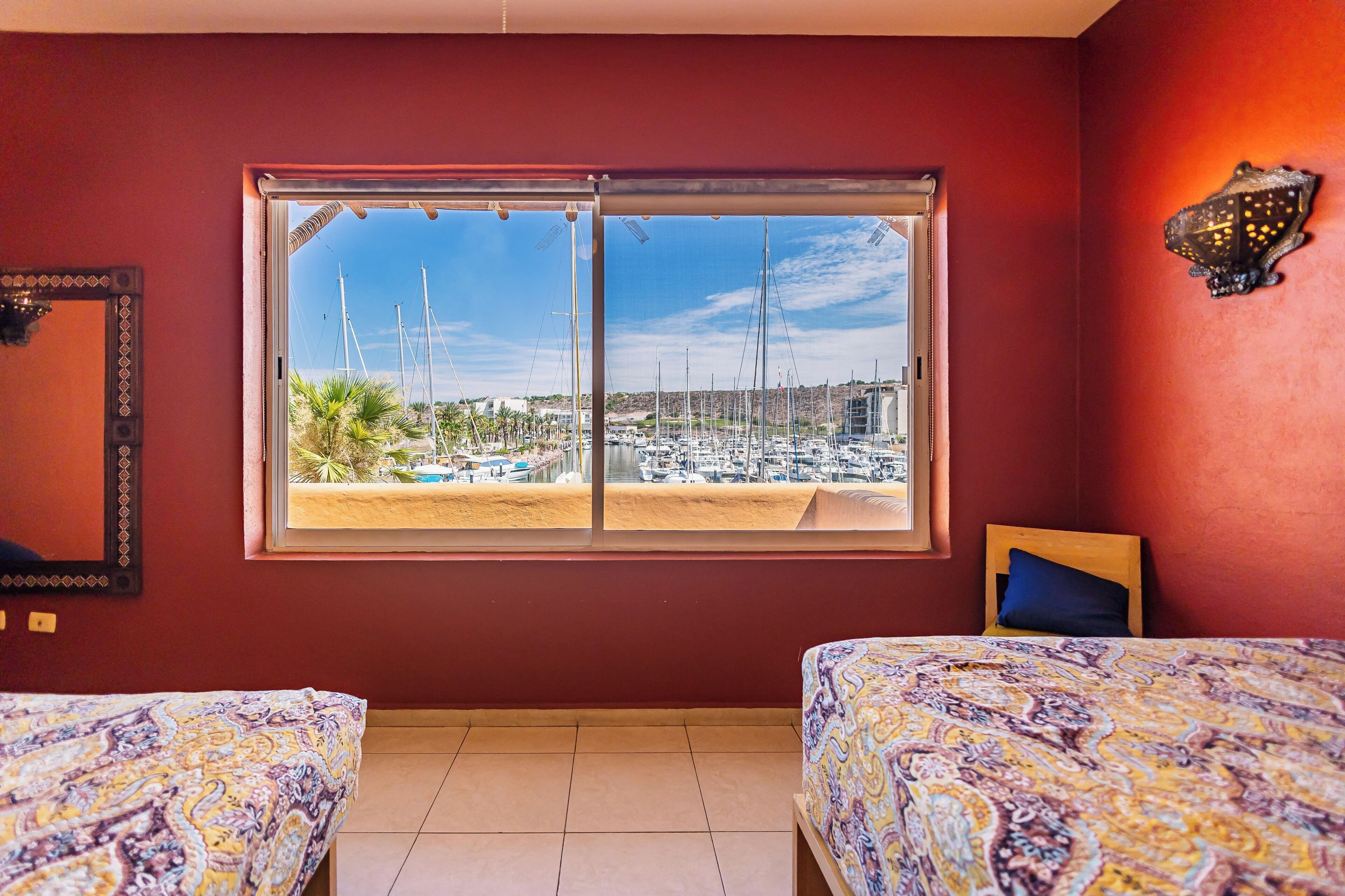 A room with two beds, a large window with a view of a marina, and a wall-mounted light fixture.