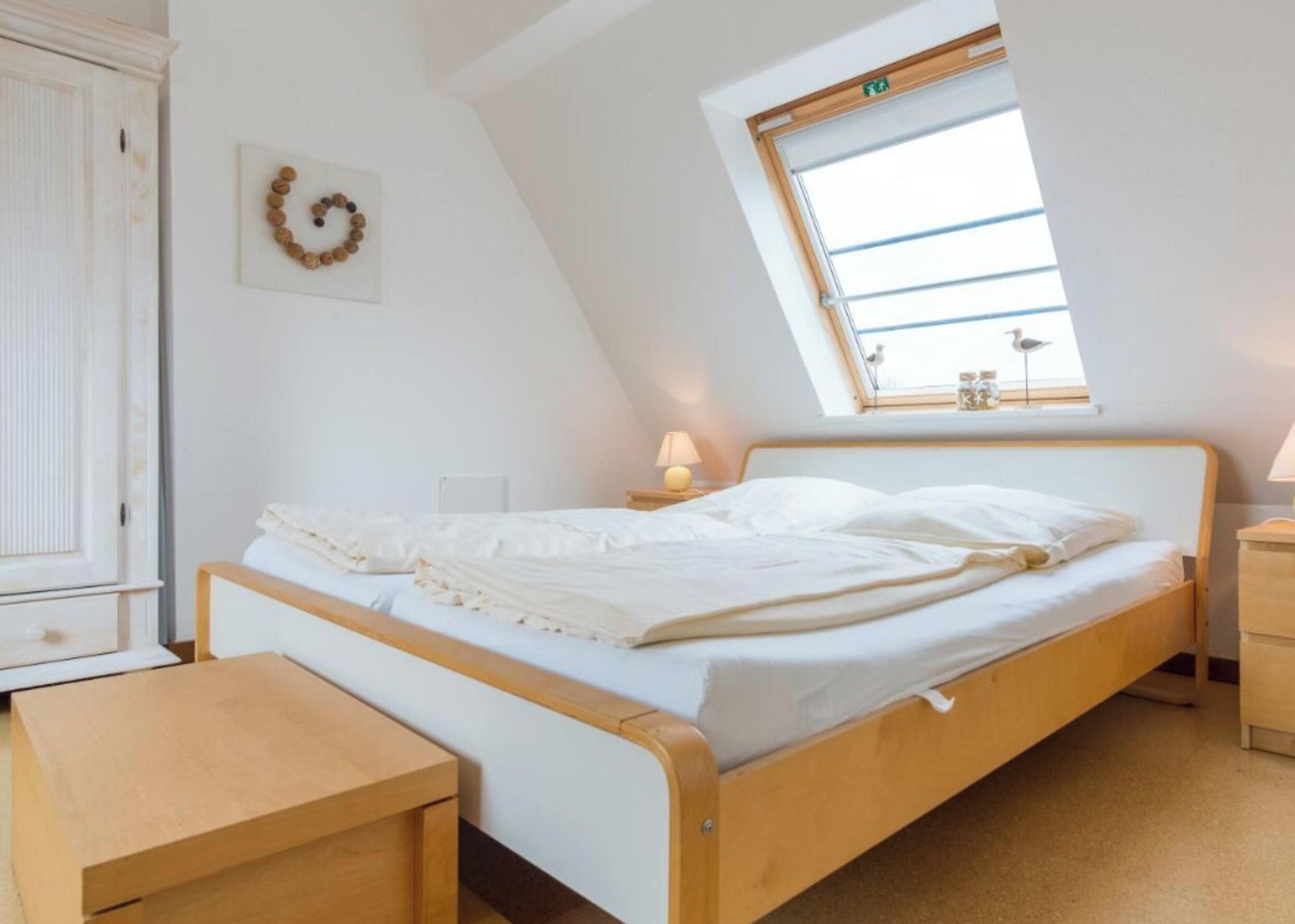 Ein Schlafzimmer mit schrägem Dach, einem Bett mit weißer Bettwäsche, einem hölzernen Nachttisch, einem kleinen Tisch und einem Fenster mit Blick auf den Himmel.