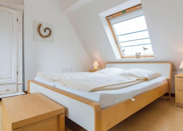 Une chambre avec un plafond en pente, un lit avec une literie blanche, une table de chevet en bois, une petite table et une fenêtre donnant sur le ciel.