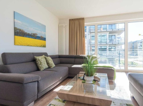A modern living room with a grey sofa, a glass coffee table, and a painting of a landscape. - Holiday Apartment With 1 Bedroom (Heiligenhafen)