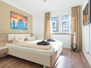 A hotel room with a bed, a bedside table, a window with curtains, a wall-mounted TV, and a painting of a sunset. - Holiday Apartment With 1 Bedroom (Heiligenhafen)