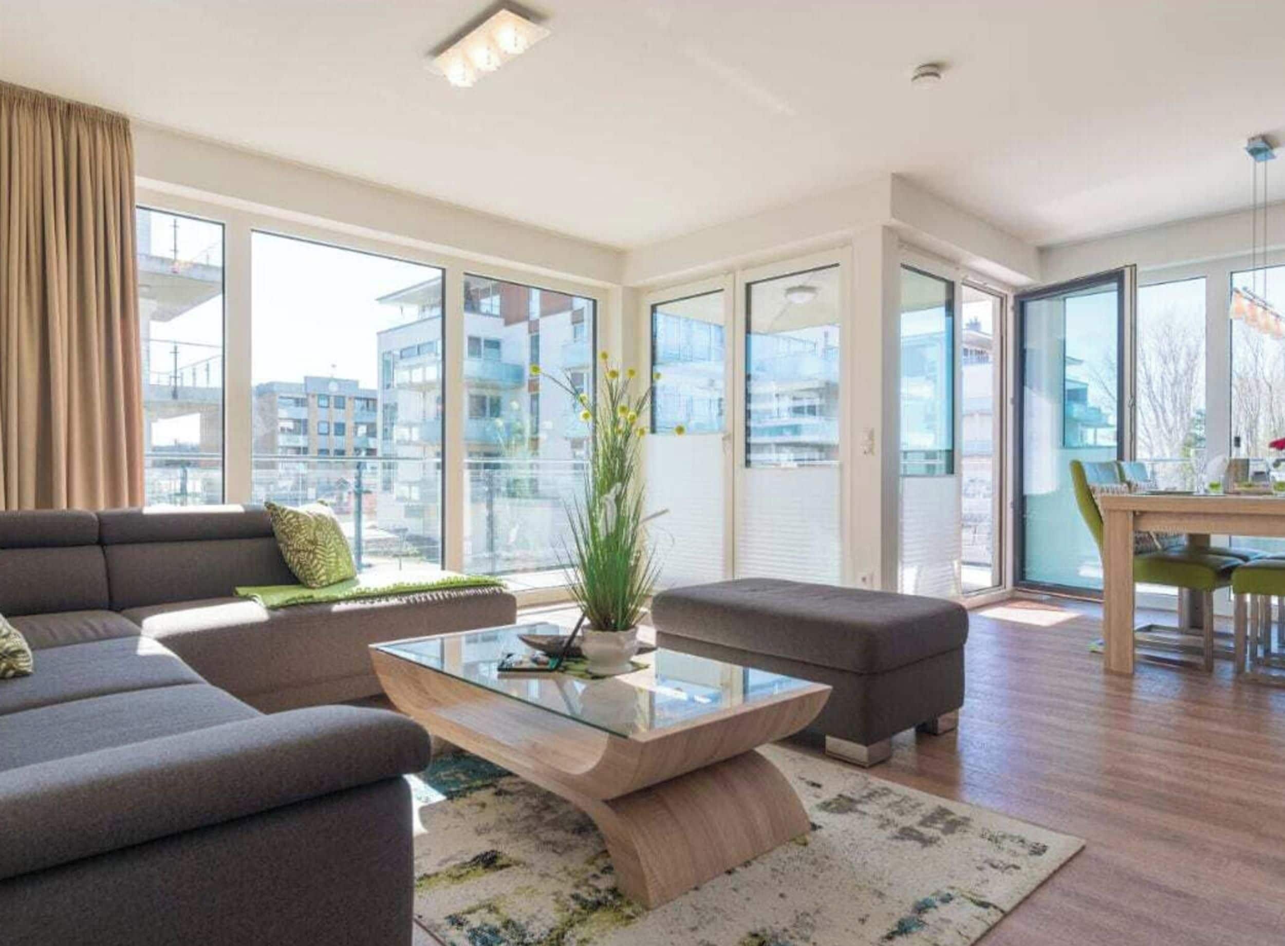 Una sala de estar moderna con sofá, reposapiés y mesa de centro de vidrio, ventanas amplias y vistas a edificios.