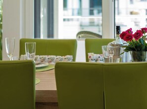 A dining area with green upholstered chairs, a wooden table, and a vase with red flowers. - Holiday Apartment With 1 Bedroom (Heiligenhafen)