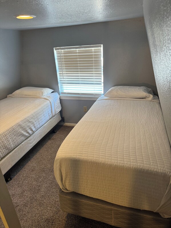 A small, single-room with two beds, a window with blinds, and a carpeted floor.