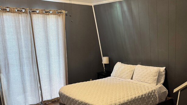 Un dormitorio con una cama, una mesita de noche, una lámpara y un ventanal con cortinas.