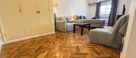 A room with a herringbone wooden floor, a sofa set, a chair, and a lamp.
