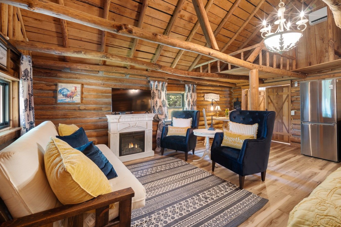 A log cabin interior with a fireplace, a television, a chandelier, and a refrigerator.