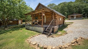 Uma cabana de toras rústica com varanda, entrada por um caminho de cascalho e área gramada ao redor.