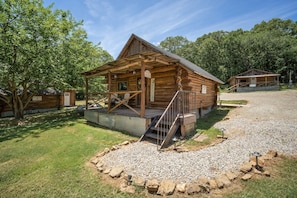 A rustic log cabin with a porch, a gravel driveway, and a surrounding grassy area. - 4 cabins-Up to 8 Guests-Unique Rural Setting but close to town! (Rudy)